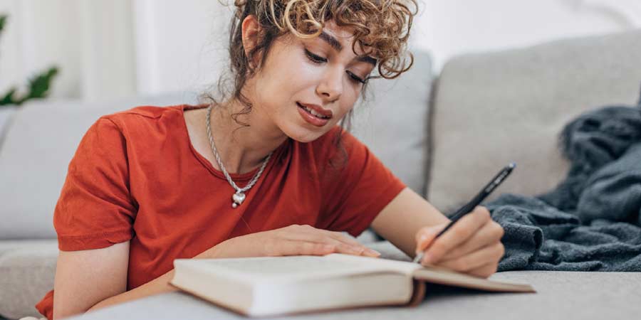 Attractive woman with curly red hair wearing a red shirt sitting down with a notepad writing out a sex list 