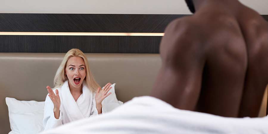 Blonde woman wearing a robe sitting in bed looking shocked as her male partner removes his robe 